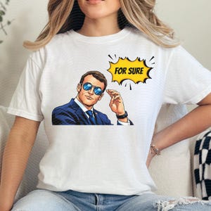 以下が含まれることがあります： 白いTシャツ。ポップアート風のイラストで、スーツとサングラス姿の男性が描かれています。黄色い吹き出しには「FOR SURE」の文字。男性は自信に満ちた表情をしています。