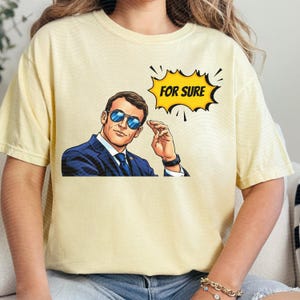 以下が含まれることがあります： サングラスをかけた男性のポップアートスタイルのイラストが特徴の、薄い黄色のTシャツ。「FOR SURE」というテキストが黄色の吹き出しに入っています。男性は青いスーツとネクタイを着ています。
