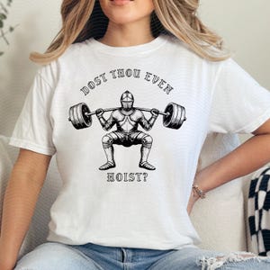 May include: White t-shirt with a black graphic of a knight lifting weights. The text "DOST THOU EVEN" arches above the knight, and "HOIST?" is below. The design is centered on the shirt.