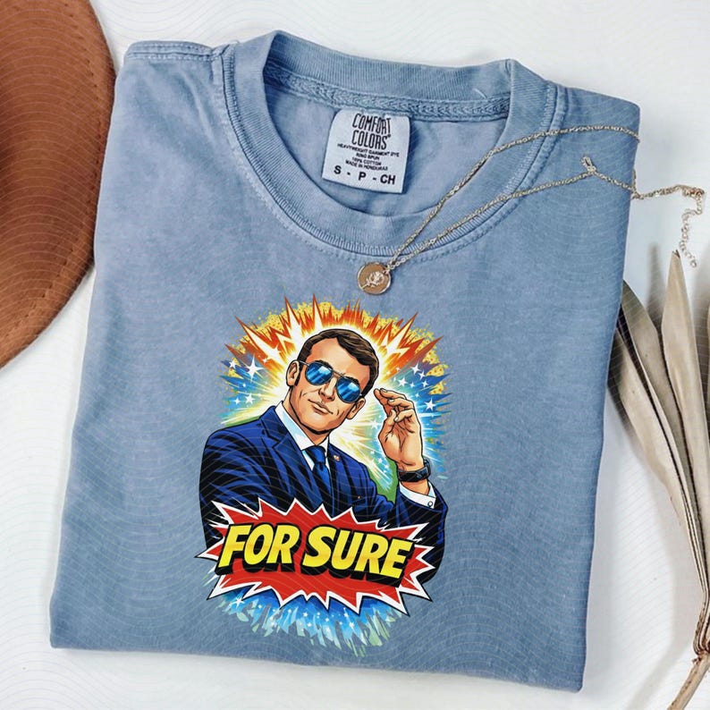 以下が含まれることがあります： サングラスをかけた男性のグラフィックと「FOR SURE」の文字がコミック風にプリントされた、ライトブルーのComfort Colors Tシャツ。ペンダント付きのゴールドネックレスがシャツの上にあります。