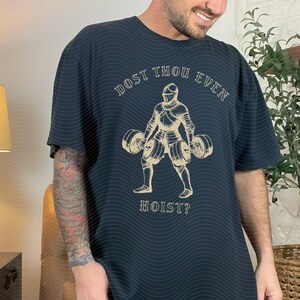 May include: Dark navy blue t-shirt with a tan illustration of a knight lifting weights. The text "DOST THOU EVEN HOIST?" is arched above and below the knight. The shirt has a relaxed fit.