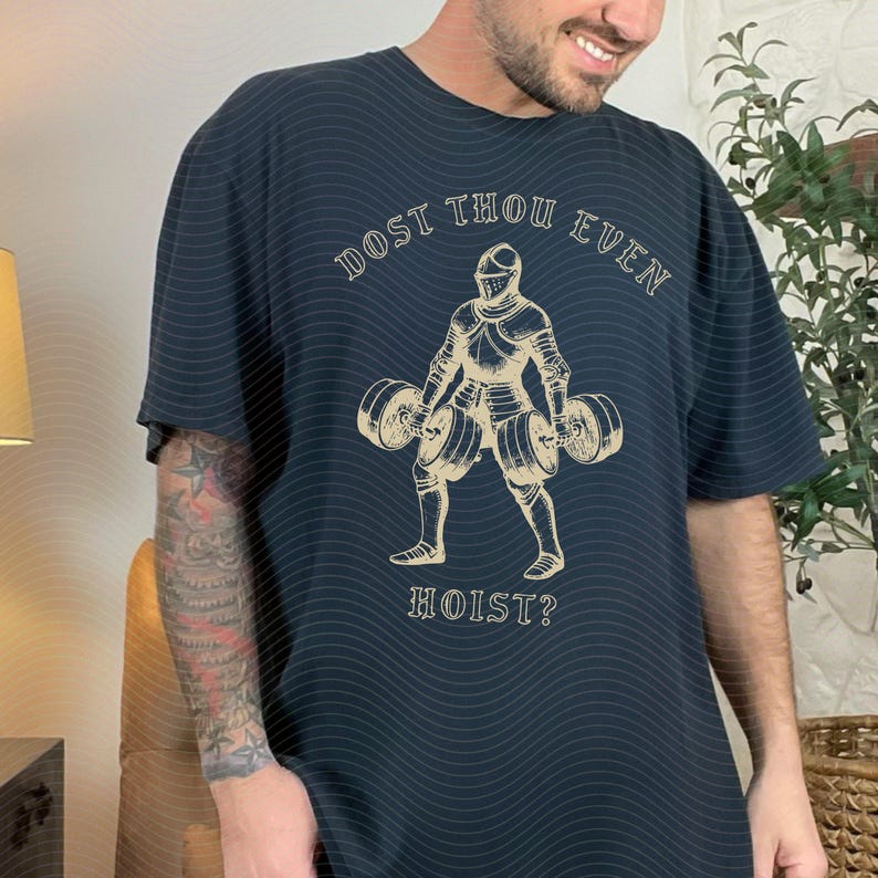May include: Dark navy blue t-shirt with a tan illustration of a knight lifting weights. The text "DOST THOU EVEN HOIST?" is arched above and below the knight. The shirt has a relaxed fit.