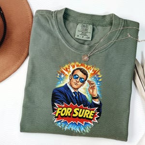 以下が含まれることがあります： サングラスをかけた男性のグラフィックと「FOR SURE」の文字が入ったオリーブグリーンのTシャツ。 Tシャツはクルーネックと半袖です。 シルバーのネックレスがTシャツの上にかけられています。 Comfort Colorsブランド。