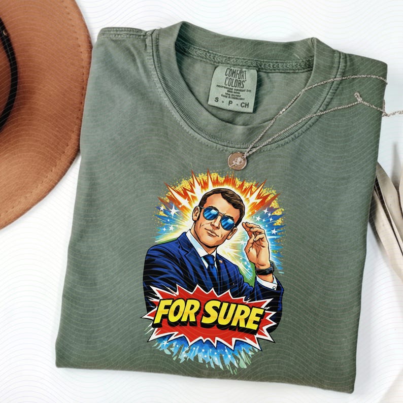 以下が含まれることがあります： サングラスをかけた男性のグラフィックと「FOR SURE」の文字が入ったオリーブグリーンのTシャツ。 Tシャツはクルーネックと半袖です。 シルバーのネックレスがTシャツの上にかけられています。 Comfort Colorsブランド。