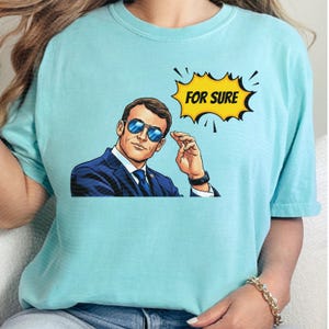 以下が含まれることがあります： サングラス、スーツ、ネクタイ姿の男性のポップアート風イラストが描かれた水色のTシャツ。「FOR SURE」と書かれた黄色の吹き出しが男性の頭上にあります。