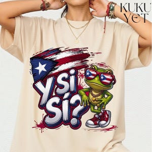 Può includere: Una t-shirt beige con la grafica di una rana verde che indossa occhiali da sole con il motivo della bandiera portoricana. Il design include una bandiera, una catena d'oro, scarpe da ginnastica rosse e il testo Y SI SÍ? in lettere bianche e blu.