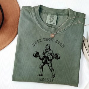 May include: Sage green t-shirt with a black graphic of a knight lifting weights, and the text "DOST THOU EVEN HOIST?". The shirt has a crew neck and short sleeves. A silver necklace with a pendant is draped across the shirt.