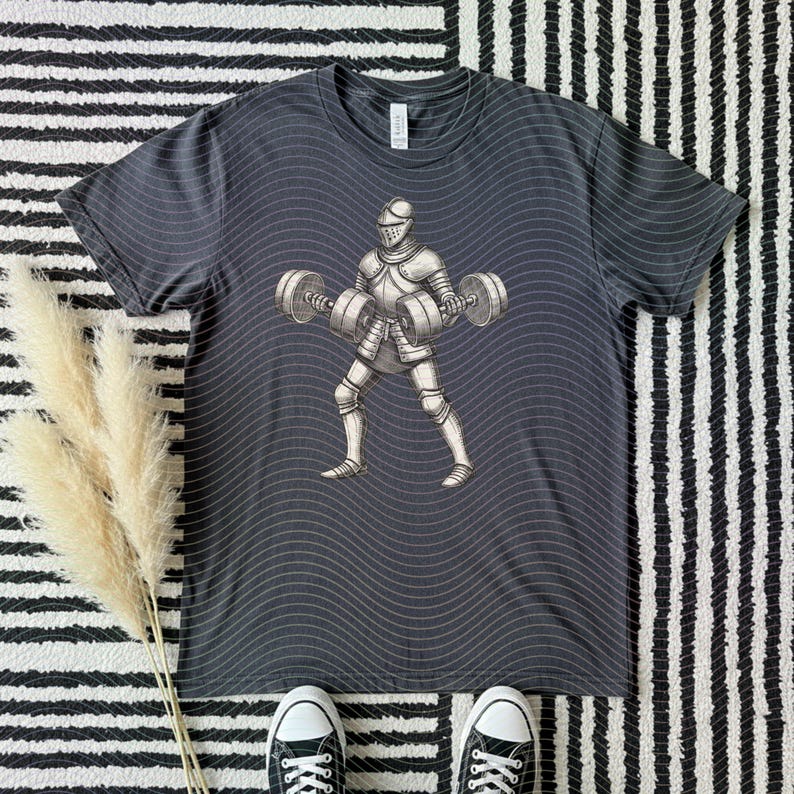 May include: Dark gray t-shirt featuring a graphic of a knight lifting barbells. The knight is wearing silver armor and a helmet. The shirt has a wavy pattern and is on a black and white striped surface.