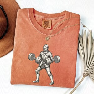 May include: A coral Comfort Colors t-shirt featuring a graphic of a knight lifting barbells. The knight is in full armor, and the barbells have weights on each end. A gold necklace and a Comfort Colors tag are visible.
