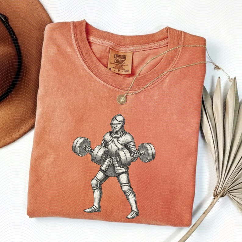 May include: A coral Comfort Colors t-shirt featuring a graphic of a knight lifting barbells. The knight is in full armor, and the barbells have weights on each end. A gold necklace and a Comfort Colors tag are visible.