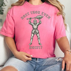May include: Pink t-shirt with the text "DOST THOU EVEN HOIST?" above and below a detailed illustration of a knight in full armor holding a mace. The shirt is a solid pink color.