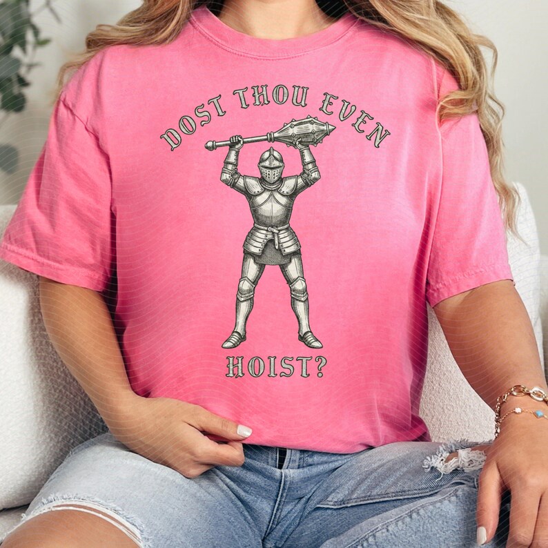 May include: Pink t-shirt with the text "DOST THOU EVEN HOIST?" above and below a detailed illustration of a knight in full armor holding a mace. The shirt is a solid pink color.
