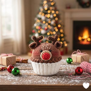 May include: A handmade crochet reindeer cupcake with a brown head, antlers, and a red nose. The cupcake has a white base and is decorated with black eyes and a white stitch pattern. Christmas ornaments and gifts surround the reindeer.