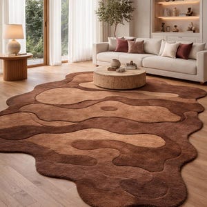 Op de afbeelding: Een groot, abstract vloerkleed in bruin-, beige- en lichtbruine tinten. Het vloerkleed heeft een organische, vloeiende vorm en ligt op een houten vloer. De kamer heeft een witte bank, een ronde salontafel en een lamp.