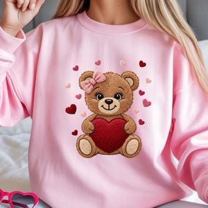 Może przedstawiać: R&oacute;żowa bluza z nadrukiem misia trzymającego czerwone serce. Miś ma r&oacute;żową kokardę i jest otoczony sercami. Bluza jest w jednolitym r&oacute;żowym kolorze, idealna na Walentynki lub na co dzień.