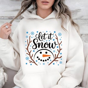 May include: Cream-colored hoodie with the text "Let it Snow" in black script, featuring a snowman face with a carrot nose and twig antlers. Blue snowflakes and dots surround the design. A cozy winter-themed sweatshirt.