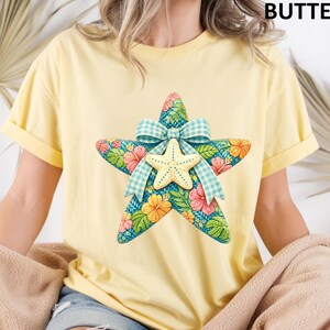 May include: A yellow t-shirt featuring a large, colorful starfish design. The starfish is decorated with floral patterns, a smaller starfish, and a blue gingham bow. The word "BUTTER" is printed at the top right.