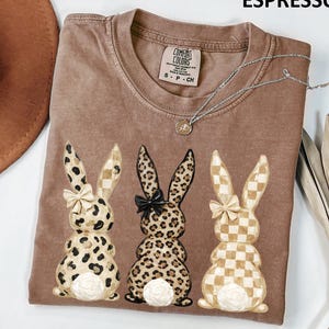 May include: Espresso-colored t-shirt with three bunny illustrations. Each bunny has a different pattern: leopard print, checkered, and a mix of both. The shirt has a crew neck and a silver necklace.