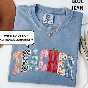 May include: Blue jean-colored t-shirt with the word "TEACHER" in a colorful, printed design. The shirt has a crew neck and short sleeves. A gold necklace with a pendant is draped across the shirt.