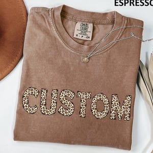 May include: Espresso-colored t-shirt with the word "CUSTOM" in leopard print across the front. The shirt has a crew neck and short sleeves. A silver necklace with a pendant is draped across the shirt. The shirt is made by Comfort Colors.
