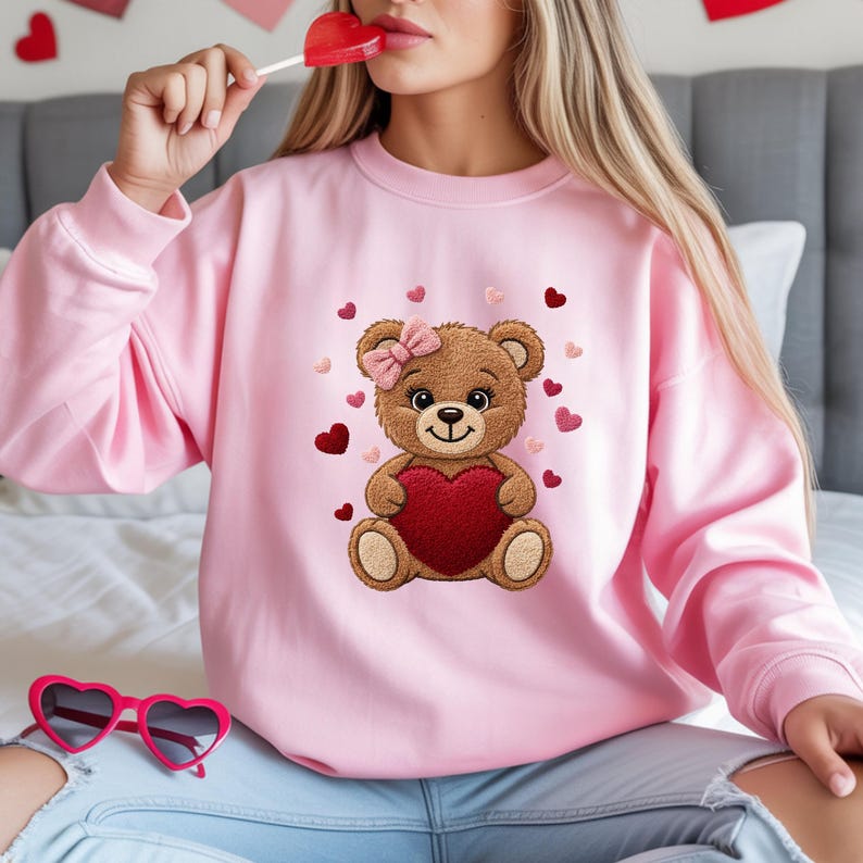 Może przedstawiać: R&oacute;żowa bluza z nadrukiem misia trzymającego czerwone serce. Miś ma r&oacute;żową kokardę i jest otoczony sercami. Bluza jest w jednolitym r&oacute;żowym kolorze, idealna na Walentynki lub na co dzień.