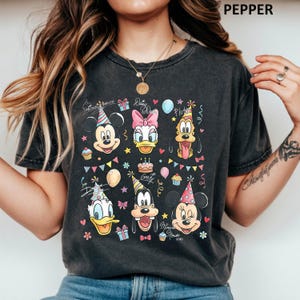Puede incluir: Camiseta gris oscuro con un gr&aacute;fico colorido de Mickey Mouse, Daisy Duck, Pluto, Donald Duck, Goofy y Minnie Mouse, todos con gorros de fiesta. El dise&ntilde;o incluye pasteles de cumplea&ntilde;os, globos y el texto "Mickey Mouse" y "Daisy Duck".