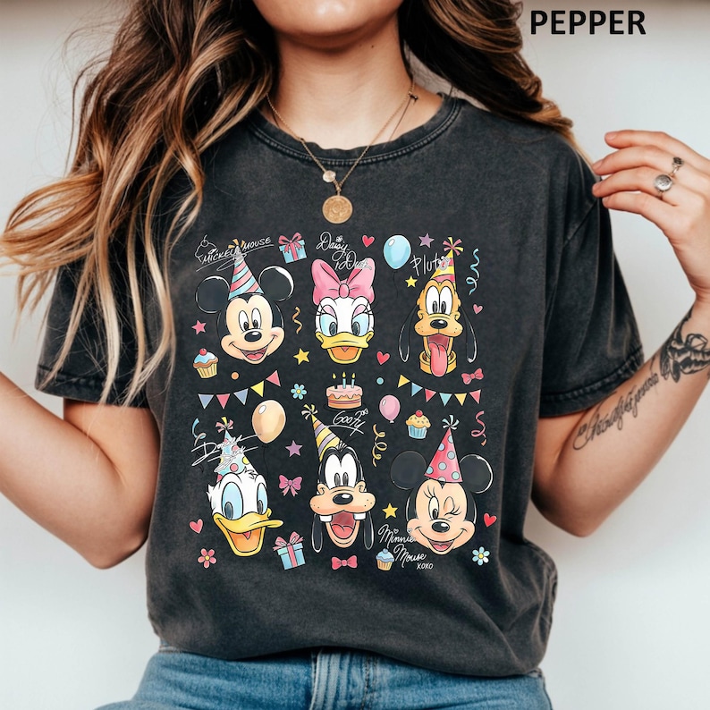Puede incluir: Camiseta gris oscuro con un gr&aacute;fico colorido de Mickey Mouse, Daisy Duck, Pluto, Donald Duck, Goofy y Minnie Mouse, todos con gorros de fiesta. El dise&ntilde;o incluye pasteles de cumplea&ntilde;os, globos y el texto "Mickey Mouse" y "Daisy Duck".