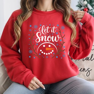 May include: Red sweatshirt with the text "Let it Snow" in white script, surrounded by a wreath and snowflakes. A snowman face with an orange carrot nose is below the text. A festive winter holiday theme.
