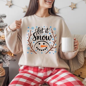 May include: Beige sweatshirt with the text "Let it Snow" in black script, featuring a snowman face with a carrot nose and twig antlers. Blue snowflakes surround the text. The person is wearing red and white plaid pajama pants.