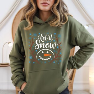 May include: An olive green hoodie with a graphic that says "Let it Snow." The design includes a snowman face with an orange carrot nose, blue snowflakes, and brown branches. The hoodie has a front pocket and a hood.