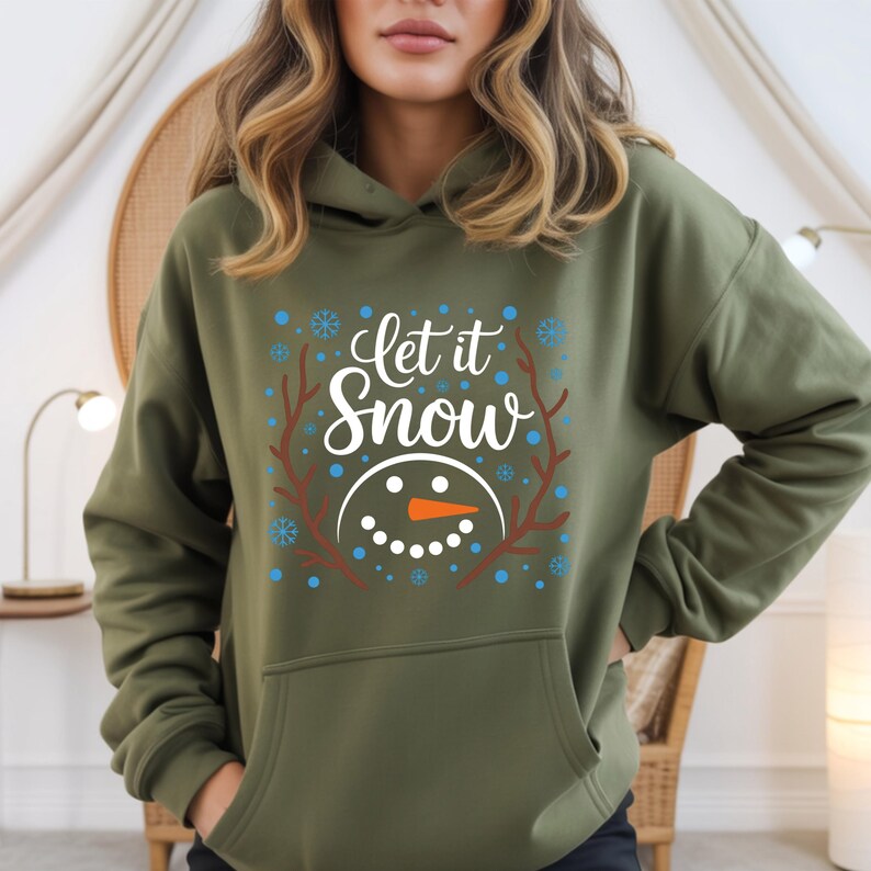 May include: An olive green hoodie with a graphic that says "Let it Snow." The design includes a snowman face with an orange carrot nose, blue snowflakes, and brown branches. The hoodie has a front pocket and a hood.