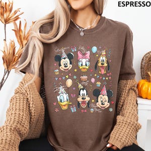 Puede incluir: Camiseta color espresso con un gr&aacute;fico colorido de Mickey Mouse, Daisy Duck, Pluto, Donald Duck, Goofy y Minnie Mouse con sombreros de fiesta. El dise&ntilde;o incluye globos, cupcakes y el texto "Mickey Mouse" y "XOXO".