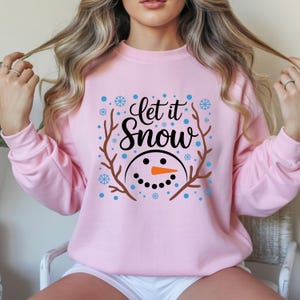 May include: A light pink sweatshirt with the phrase "Let it Snow" in black script, surrounded by a snowman face and snowflakes. The snowman has a carrot nose and a smiling mouth. Brown branches frame the design.