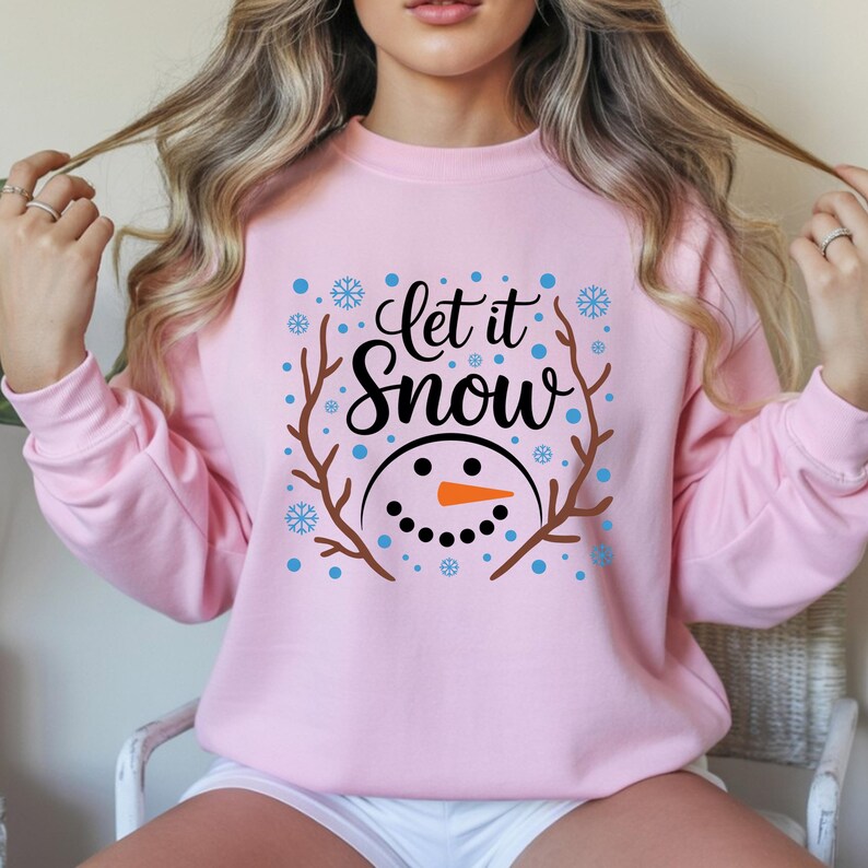 May include: A light pink sweatshirt with the phrase "Let it Snow" in black script, surrounded by a snowman face and snowflakes. The snowman has a carrot nose and a smiling mouth. Brown branches frame the design.