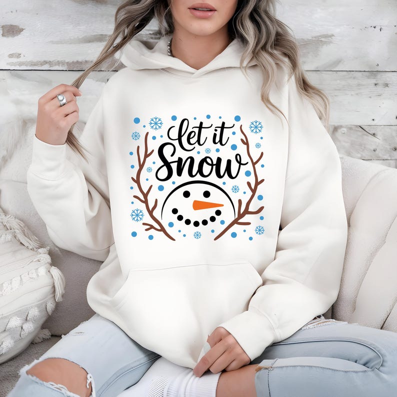 May include: Cream-colored hoodie with the text "Let it Snow" in black script, featuring a snowman face with a carrot nose and twig antlers. Blue snowflakes and dots surround the design. A cozy winter-themed sweatshirt.