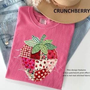 May include: Pink t-shirt with a strawberry patchwork design. The strawberry features various patterns, including gingham, floral, and hearts. The text "CRUNCHBERRY" is above the shirt. The design is a faux patchwork print effect.