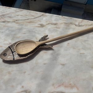 May include: A light-coloured ceramic spoon rest shaped like a fish with black hand-painted outlines and a circular eye detail. A smooth, light-brown wooden spoon rests horizontally across the centre of the fish-shaped ceramic dish.