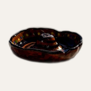 Puede incluir: Un plato de cerámica vidriada marrón con forma de sombrero, con una corona central elevada y un ala ancha. El borde exterior del ala presenta una serie de pequeños puntos pintados de colores azul, amarillo y verde sobre el acabado marrón.