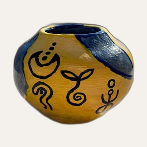 Peut inclure: Un petit pot en céramique rond peint à la main, doté d'une base jaune vif avec des taches bleu foncé contrastantes. La surface est décorée de symboles noirs dessinés à la main, dont un croissant de lune, une plante en spirale et une silhouette assise.