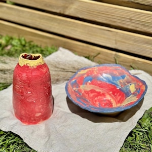 Pode incluir: Um vaso de cerâmica e uma tigela a condizer com um esmalte vibrante e multicolorido. O vaso é principalmente vermelho com uma borda amarela, enquanto a tigela apresenta um padrão de redemoinho em vermelho, azul e amarelo com uma borda ondulada.