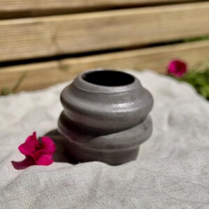 Puede incluir: Un pequeño jarrón de cerámica gris oscuro con una forma redondeada de varios niveles que presenta dos crestas horizontales prominentes alrededor del cuerpo. El jarrón tiene un acabado liso de aspecto metálico y una amplia abertura circular.