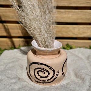 Pode incluir: Um pequeno vaso de cerâmica redondo na cor pêssego com uma borda branca larga e alargada. O exterior apresenta desenhos em espiral castanho-escuros pintados à mão. Um conjunto de hastes de erva-das-pampas bege seca está dentro do vaso.