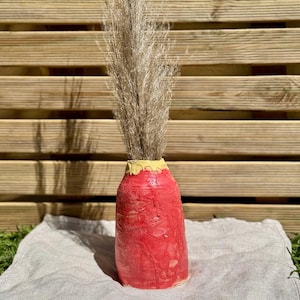 Può includere: Un vaso cilindrico in ceramica con un esterno testurizzato rosso brillante e un bordo giallo sulla parte superiore. Il vaso contiene una composizione di erba beige secca, alta e piumosa che si estende verticalmente dall'apertura del vaso.