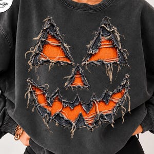 Könnte beinhalten: Ein anthrazitfarbenes Sweatshirt mit Rundhalsausschnitt und einem abgenutzten, ausgefransten Kürbisgesicht-Motiv. Die Augen, die Nase und der gezackte Mund des Kürbisses sind in leuchtendem Orange mit rauen Stoffkanten für einen Textureffekt.