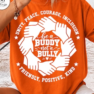 May include: An orange t-shirt featuring a white graphic of hands joined in a circle. The text reads: UNITY, PEACE, COURAGE, INCLUSION, be a BUDDY not a BULLY, FRIENDLY, POSITIVE, KIND. The design includes stars and a small heart.