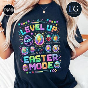 以下が含まれることがあります： 「LEVEL UP」と「EASTER MODE」の文字が入った、カラフルなグラフィックデザインの黒いTシャツ。ピクセル化された卵、稲妻、ゲームのような要素が含まれています。Tシャツはクルーネックです。