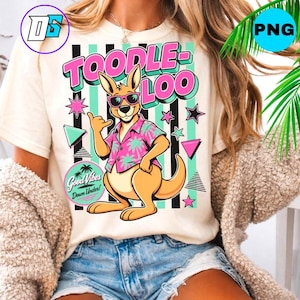 Op de afbeelding: Crèmekleurig T-shirt met een cartoon kangoeroe met een zonnebril en een Hawaiiaans shirt. Het shirt heeft de tekst "TOODLE-LOO" in roze en een "Good Vibes Down Under!" afbeelding. Het ontwerp bevat sterren en geometrische vormen.