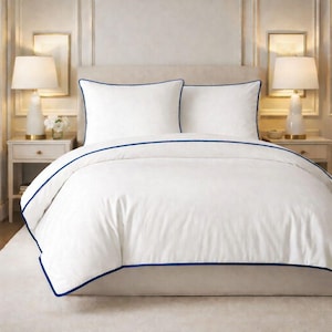 Peut inclure: Ensemble de literie blanc avec une bordure bleu marine. L'ensemble comprend une housse de couette et deux taies d'oreiller. La literie est sur un lit avec deux tables de chevet blanches, chacune avec une lampe.