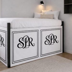Könnte beinhalten: Ein weißer Bettvolant für ein Hochbett mit einem schwarzen Doppellinienrand und einem großen schwarzen Monogramm mit den Buchstaben SPR in einer dekorativen Schreibschrift. Die Stoffbahnen sind am schwarzen Metallrahmen des Bettes befestigt.