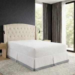 May include: A beige upholstered bed with a buttoned headboard and a white bed skirt with a grey border. The bed is in a well-lit room with a large window and dark curtains. A neutral-toned rug is on the floor.